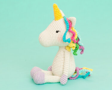 Cream crocheted amigurumi unicorn with yellow horn and pastel curly mane sitting on a mint background