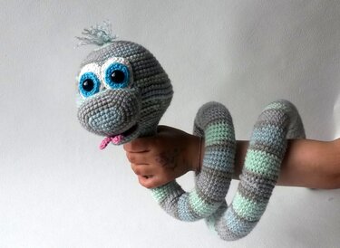 Gehäkelte, geringelte Amigurumi‑Schlange in Grau, Hellblau und Mintgrün mit großen blauen Augen und rosa Zunge, um einen Arm gewickelt.