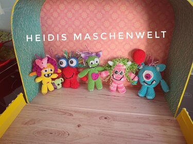 Sechs bunte gehäkelte Amigurumi-Monster mit unterschiedlichen Gesichtern und Aufhängeschlaufen auf einer Holzfläche