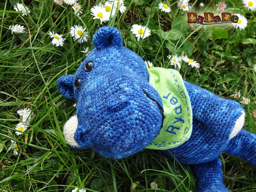 Blaues gehäkeltes Nilpferd-Amigurumi mit hellgrünem Oberteil liegt in einer Wiese mit Gänseblümchen.