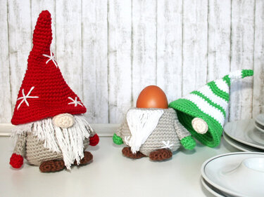 Three crocheted gnomes: one tall red-hatted gnome and two egg cozies, one holding a brown egg.