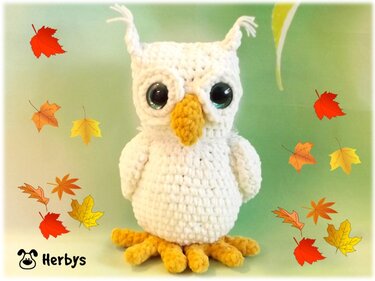 Weiße gehäkelte Amigurumi-Eule mit großen glänzenden Augen, gelbem Schnabel und gelben Füßen, umgeben von roten, orange- und gelben Herbstblättern vor grünem Hintergrund.