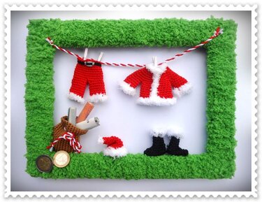 Grüner, gehäkelter Rahmen mit kleinen rot‑weißen Weihnachtsmann‑Kleidungsstücken an einer Schnur und einem Beutel mit Geldscheinen und Münzen