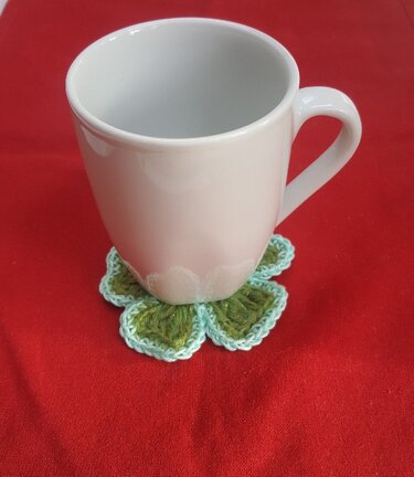 Weiße Tasse auf gehäkeltem Kleeblatt-Untersetzer in Grün und Hellblau auf rotem Stoff.