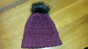 Bordeauxfarbene gestrickte Damenmütze mit Umschlag und dunklem Bommel auf Holzfläche