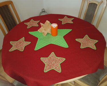 Runder Tisch mit roter Decke, großem grünen Häkelstern in der Mitte, sechs gehäkelten Stern-Untersetzern, Holzstern und orangefarbener Kerze