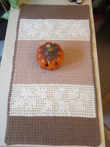 Gehäkelter Tischläufer in Dunkelbraun, Hellbraun und Creme mit durchbrochenem Herzmuster, auf einem Tisch mit Kürbisdekoration.