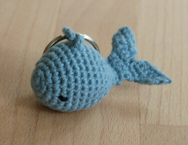 Gehäkelter hellblauer Wal mit Metall-Schlüsselring auf heller Holzoberfläche, sichtbare Häkelmaschen und kleines schwarzes Auge