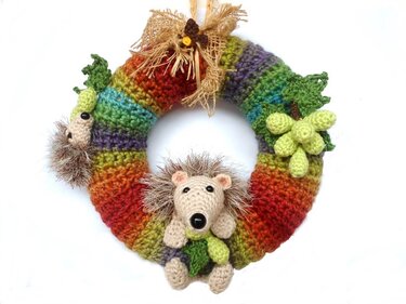 Bunter gehäkelter Kranz in Streifenoptik mit zwei Igel-Amigurumis, gehäkelten Trauben, Blättern und Juteband-Schleife auf weißem Hintergrund.
