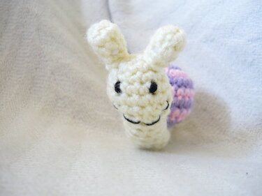 Nahaufnahme einer gehäkelten Amigurumi-Schnecke mit cremefarbenem Körper und lila-rosa gestreifter Schale