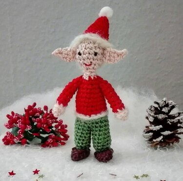 Gehäkelter Weihnachtswichtel mit roter Zipfelmütze, rotem Oberteil und grüner Hose vor weihnachtlicher Dekoration