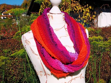Gehäkelter Loop‑Schal in Orange, Rot und Lila auf einer Puppe vor herbstlicher Gartenkulisse.