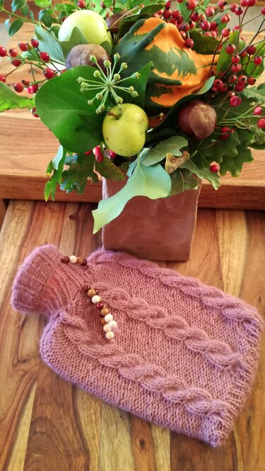 Gestrickte Wärmflaschenhülle in Rosa-Lila mit Zopfmuster und Holzperlenkette auf Holz, davor herbstliche Dekoration.
