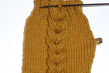 Detailaufnahme einer gestrickten senfgelben Armstulpe mit Zopfmuster, Stricknadel und Maschenhalter