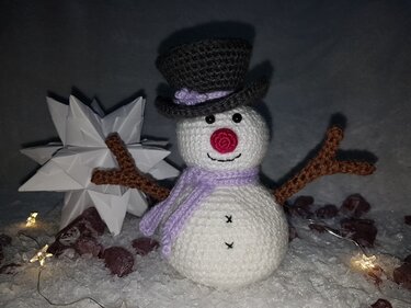 Gehäkelter Amigurumi‑Schneemann mit schwarzem Zylinder, lila Schal, roter Nase und Astarmen vor Papierstern und Lichterkette