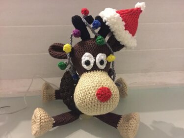 Handgehäkeltes Rentier mit braunem Körper, beiger Schnauze, roter Nase, weißen Augen, roter Weihnachtsmütze und bunter Lichterkette um das Geweih.