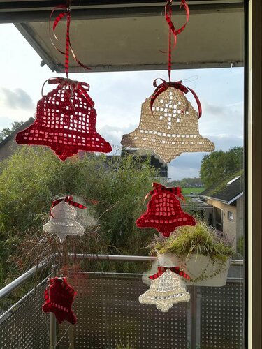 Gehäkelte Weihnachtsglocken in Rot und Creme mit roten Bändern, aufgehängt vor einem Balkonfenster.