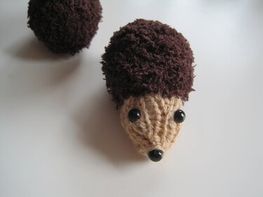 Gestrickter Bommel-Igel mit dunkelbraunem Körper und hellbraunem Gesicht, schwarze Perlenaugen und -nase