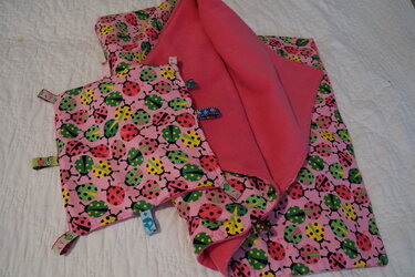 Rosa Babydecke mit Marienkäfer-Muster und kleines Tasttuch mit bunten Stofflaschen auf weißer Unterlage