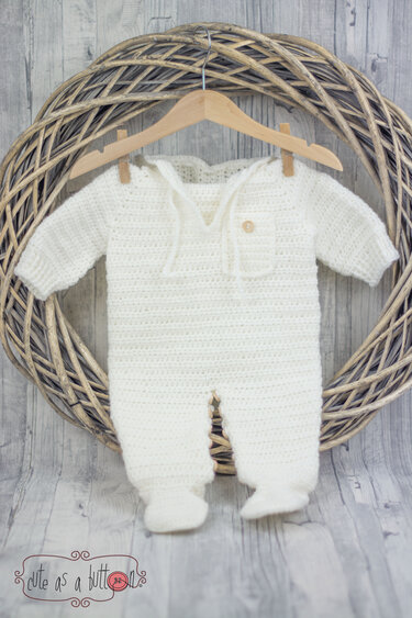 Cremefarbener gehäkelter Baby-Overall mit Füßchen an Holzbügel vor geflochtenem Kranz
