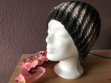 Gestrickte gestreifte Mütze in Dunkel- und Hellgrau auf weißem Schaufensterkopf, rosa Kunstorchideen daneben