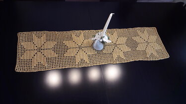 Heller gehäkelter Tischläufer mit Sternenmuster und einer weißen Kerze in Blumenarrangement auf dunklem Tisch