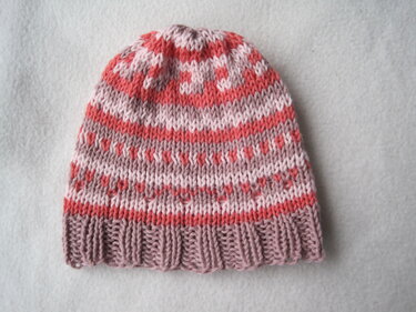 Nahaufnahme einer gestrickten Babymütze mit Herz- und Streifenmuster in Rosa, Koralle und Mauve auf hellem Hintergrund.