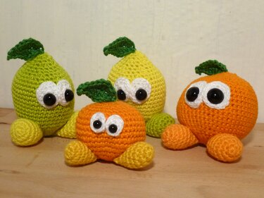 Vier gehäkelte runde Obstfiguren in Gelb und Orange mit großen weißen Augen und grünen Blättern auf einer Holzoberfläche