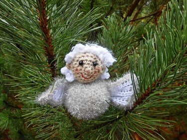 Silberweißes gehäkeltes Amigurumi-Engelchen mit flauschigem Körper und weißen Flügeln an einem Kiefernzweig
