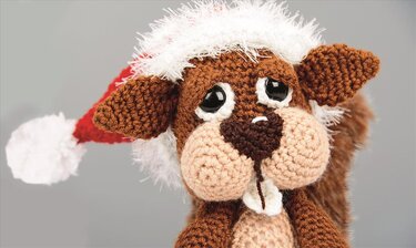 Nahaufnahme eines gehäkelten Amigurumi-Eichhörnchens in Braun mit großen Augen, Zähnchen und roter Weihnachtsmütze