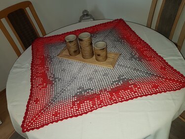 Quadratisches gehäkeltes Deckchen in Grau mit roter Umrandung auf einem runden, weiß gedeckten Tisch, darauf drei rustikale Kerzenhalter aus Holz auf einem kleinen Tablett.