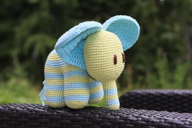 Gehäkelter Amigurumi‑Elefant in Pastellgelb und Hellblau mit Streifen, auf dunklem geflochtenem Möbelstück.