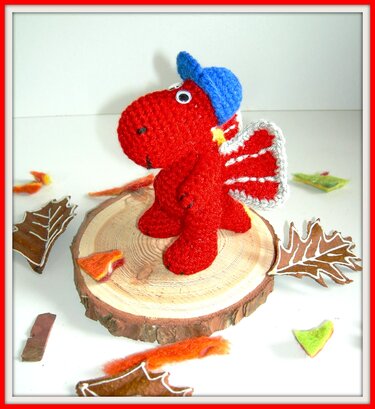 Rot gehäkelter Amigurumi-Drache mit blauer Mütze und weißen Flügelkanten auf einer Holzscheibe
