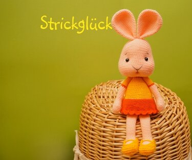 Gehäkelter Amigurumi-Hase in orange-gelbem Kleid auf einem geflochtenen Korb vor grünem Hintergrund mit dem Schriftzug 'Strickglück'.