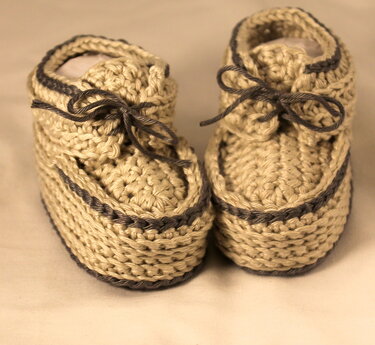 Zwei beige gehäkelte Babyschuhe mit dunkler Umrandung und Schnürbändern