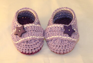 Ein Paar lila gehäkelte Babyschuhe mit weißen Streifen und dunklen Sternknöpfen von oben.