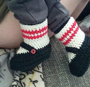 Gehäkelte Babyschuhe in Schwarz mit weiß-roten Streifen und rotem Knopf am Riemen, getragen von einem Kind.