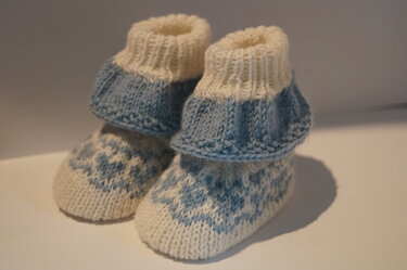 Ein Paar gestrickte Babyschuhe in Hellblau und Cremeweiß mit geometrischem Fair‑Isle‑Muster und geripptem Schaft.