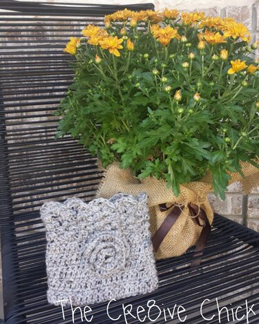 Gehäkelte graubeige Stulpe mit Rosenapplikation auf Lattenstuhl neben gelben Chrysanthemen