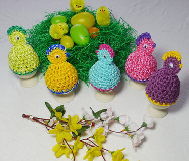 Sechs bunte gehäkelte Küken-Eierwärmer auf Eierbechern vor grünem Ostergras und bunten Deko-Eiern