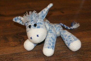 Gehäkeltes blau-weißes Amigurumi-Eselchen mit Fransenmähne und geflochtenem Schwanz auf Holzfußboden