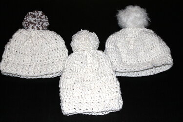 Drei weiße gehäkelte Mützen mit Bommeln auf schwarzem Hintergrund; eine Mütze hat einen braun-weißen Bommel.