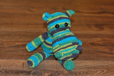 Gestreiftes Amigurumi‑Nilpferd in Blau, Grün und Grau mit schwarzen Knopfaugen auf Holzuntergrund.