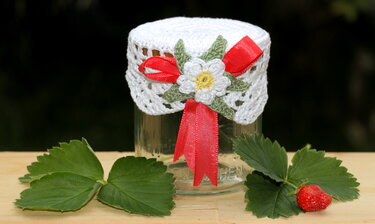 Glas mit weißer gehäkelter Deckelhaube, rotem Band und Erdbeerblüte, umgeben von Erdbeerblättern und einer Erdbeere auf Holz.