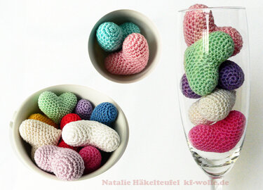 Gehäkelte Mini‑Herzen und Amigurumi‑Kugeln in Pastell- und kräftigen Tönen, präsentiert in zwei weißen Schalen und einem schlanken Glas auf weißem Hintergrund.