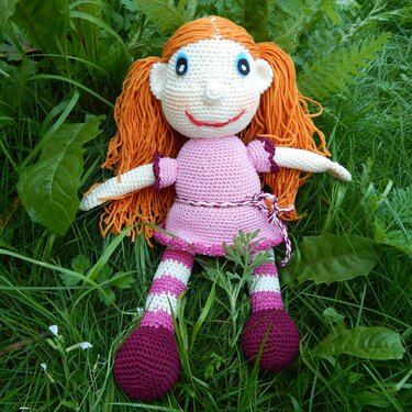 Gehäkelte Amigurumi-Puppe mit orangefarbenen Wollhaaren, rosa Häkelkleid, Ringelbeinen und dunklen Häkelschuhen im Gras.