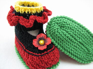 Gestrickter Babyschuh rot und schwarz mit gelbem Bündchen, roter Rüsche, grüner Sohle und Blütenapplikation