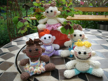 Fünf gehäkelte Amigurumi-Puppen auf einem gemusterten Gartentisch, eine in einem roten Blumentopf