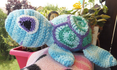 Gehäkelte Amigurumi-Schildkröte in Blau, Türkis und Grün mit violetten Details, auf einem Kissen vor Pflanzen.