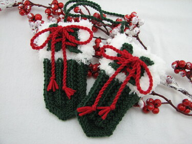 Zwei kleine gestrickte Babyfäustlinge in Grün, Weiß und Rot mit roten Schleifen.